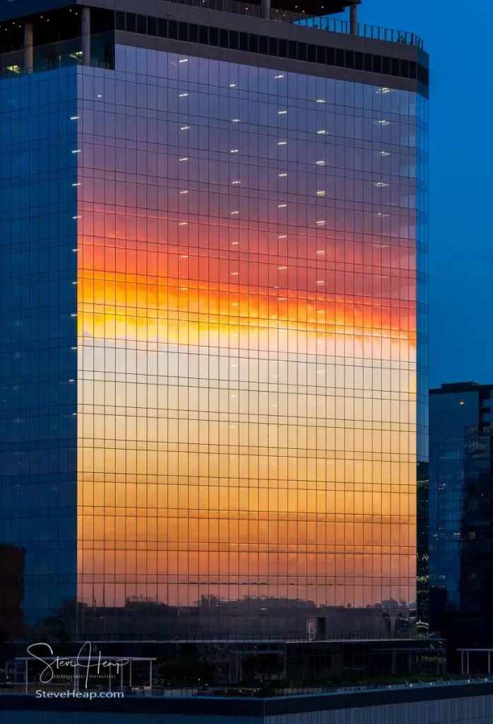 Sunset clouds reflected in glass windows on sixth and guadalupe st office and apartment building in Austin Texas. This stunning photograph captures the mesmerizing reflection of a vibrant sunset on the glass facade of the Sixth and Guadalupe building in Austin, Texas. The building's sleek, modern architecture is accentuated by the grid of reflective windows, which perfectly mirror the brilliant hues of the setting sun. The sky transitions from deep blue at the top to a fiery gradient of orange, yellow, and red, creating a breathtaking visual spectacle. The reflection is so vivid and clear that it almost appears as if the building itself is glowing with the colors of the sunset. The image is further enhanced by the subtle details of the cityscape at the bottom, adding depth and context to the scene. This photograph is not just a simple cityscape; it is a captivating blend of natural beauty and urban sophistication. It would make a striking addition to any art collection, bringing a touch of elegance and a reminder of nature's splendor to any space. The image is a testament to the photographer's skill in capturing the perfect moment, making it a must-have for anyone who appreciates fine art photography.